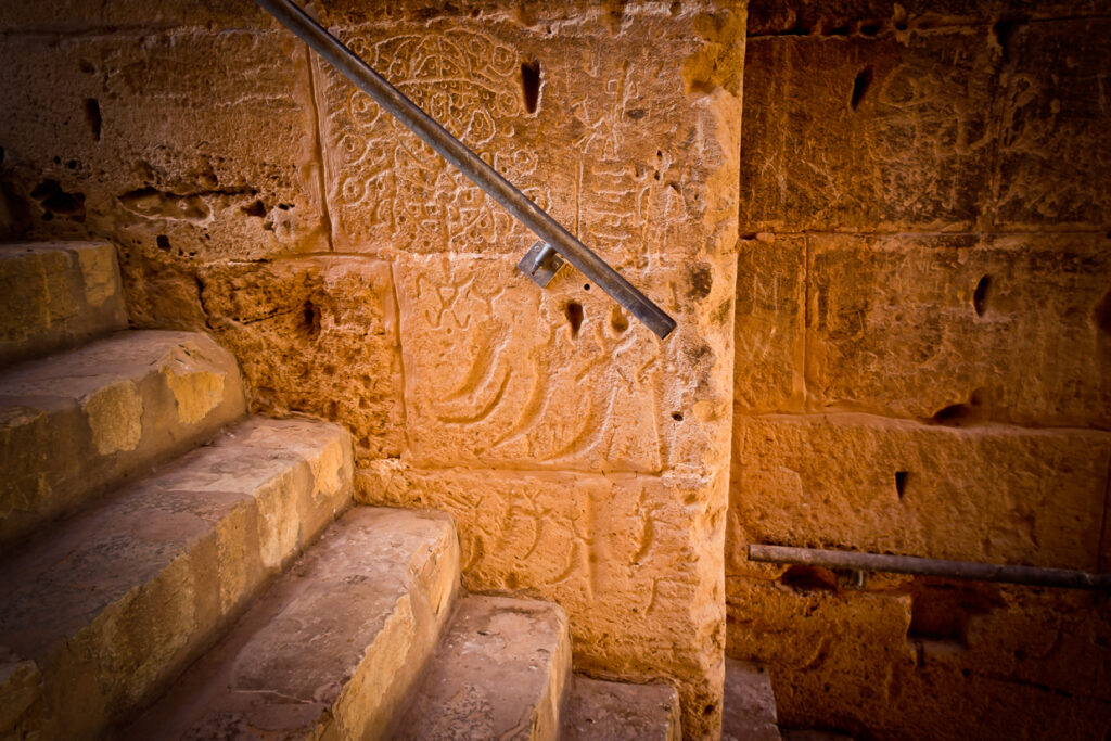 Tre pugnali curvi incisi nella pietra accanto a iscrizioni arabe e a una scala dell’anfiteatro di El Jem.